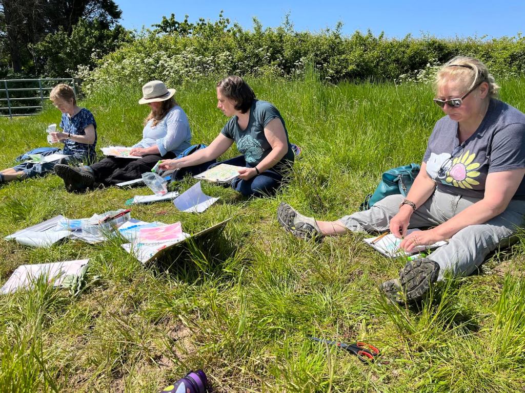 group of people sitting on the grass in the countryside with blue skies and sun shining drawing and painting